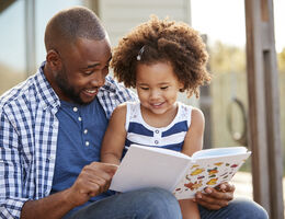 A man smiles as he reads to the smiling child on his lap.